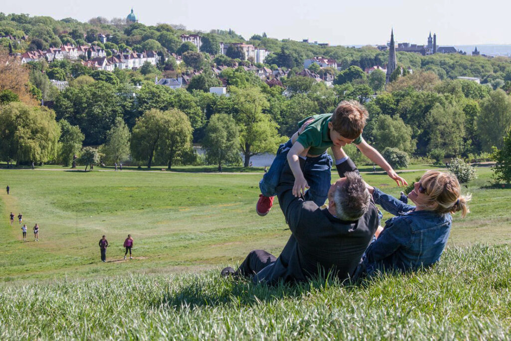 Man and woman holding toddler boy in the air on large green heath with houses on a hill in the distance